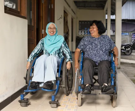 Two women in wheelchairs outside their office door, smiling and talking to each other.