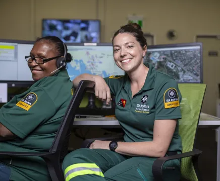 Two people sit at a desk, wearing green paramedic uniforms, they face the camera smiling. On the desk in front of them are maps and excel spreadsheets.