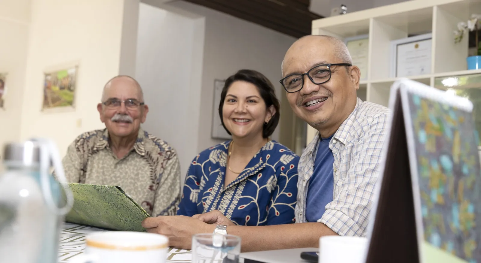 Three people are sitting at a desk in an office, laptops, cups of tea, and maps are visible. They are all smiling at the cameara.