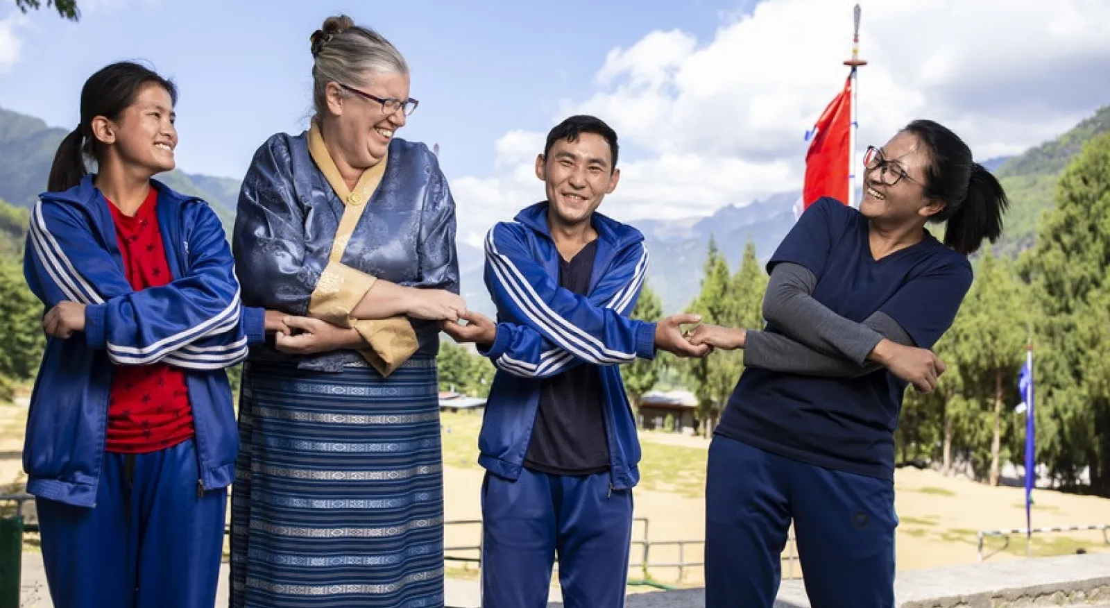 Four people in blue and red traditional Bhutanese clothing smiling and laughing while standing in front of a mountainous background.