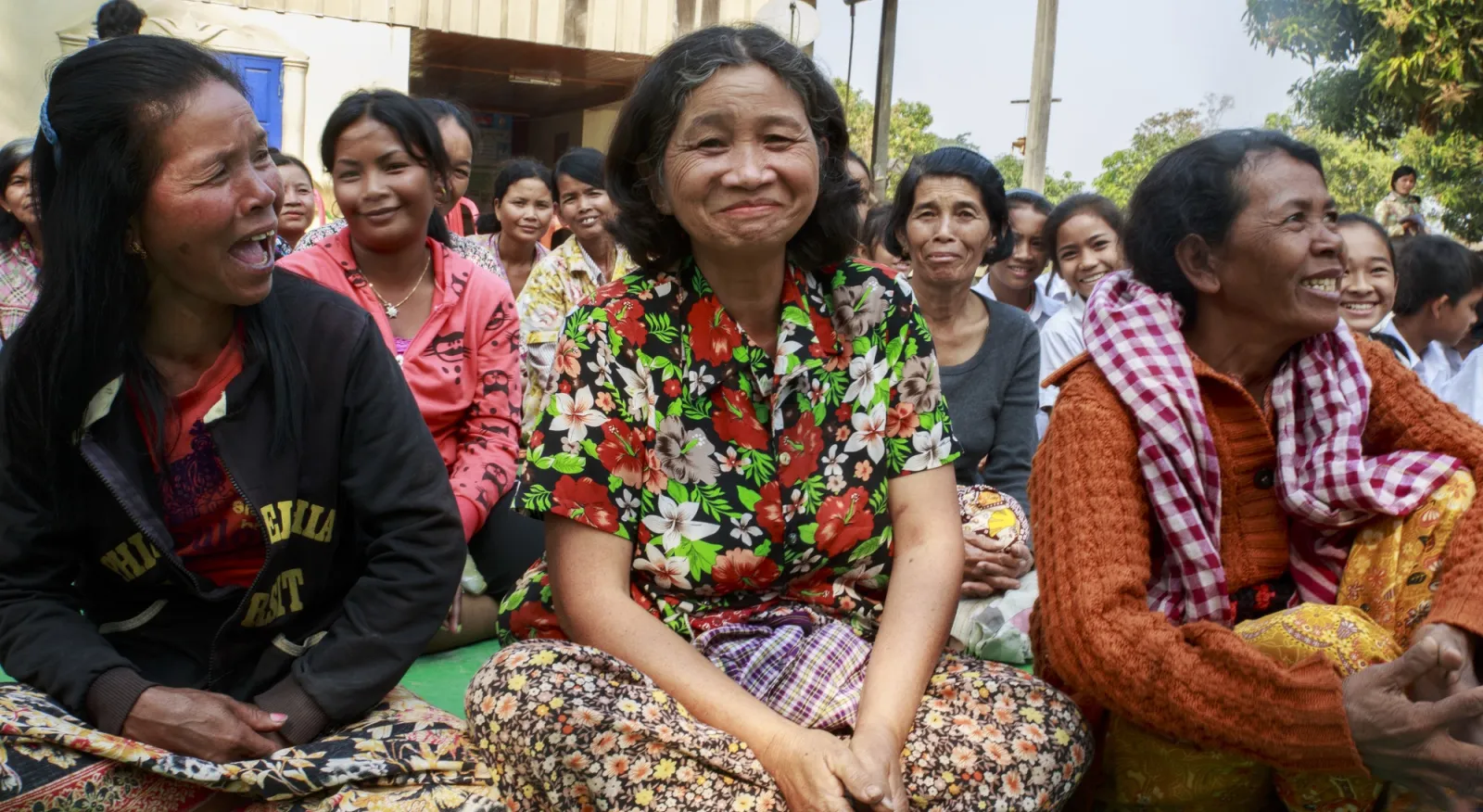 Three women sit on the ground in Cambodia, legs crossed and smiling in mid-conversation.