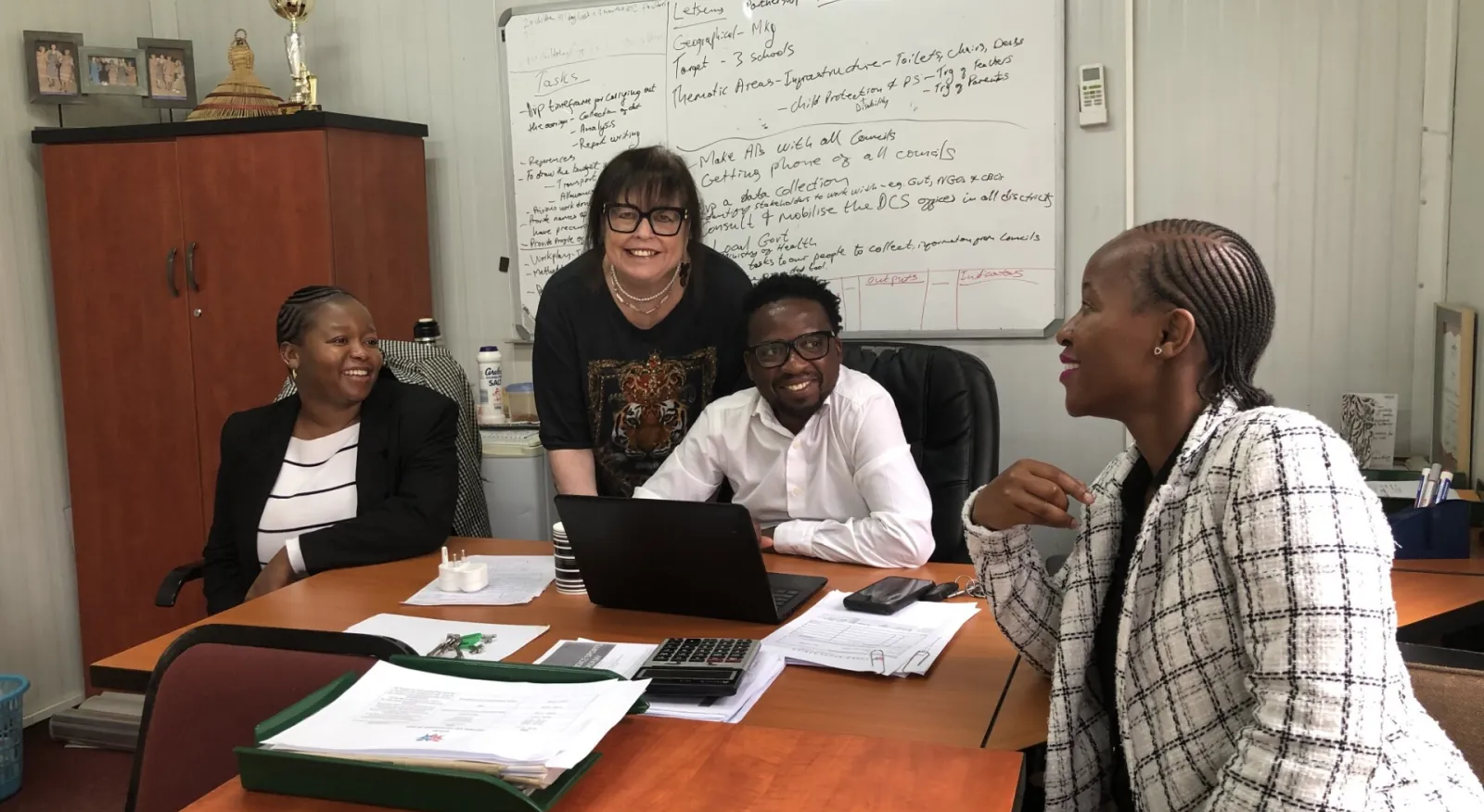 Three women and a man are sitting in a boardroom, chatting and smiling at each other. Behind them is a whiteboard full of text. In front of them on the table is papers and laptops.