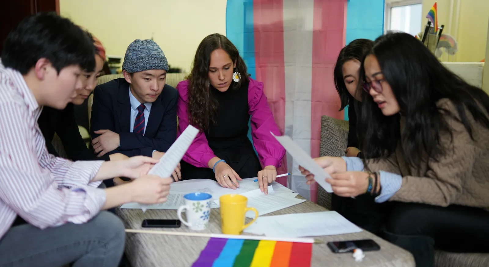 A group of young people are sitting around a small coffee table, with their heads down looking over papers. They are surrounded by a LGBTIQA+ flag and the Trans Right flag.