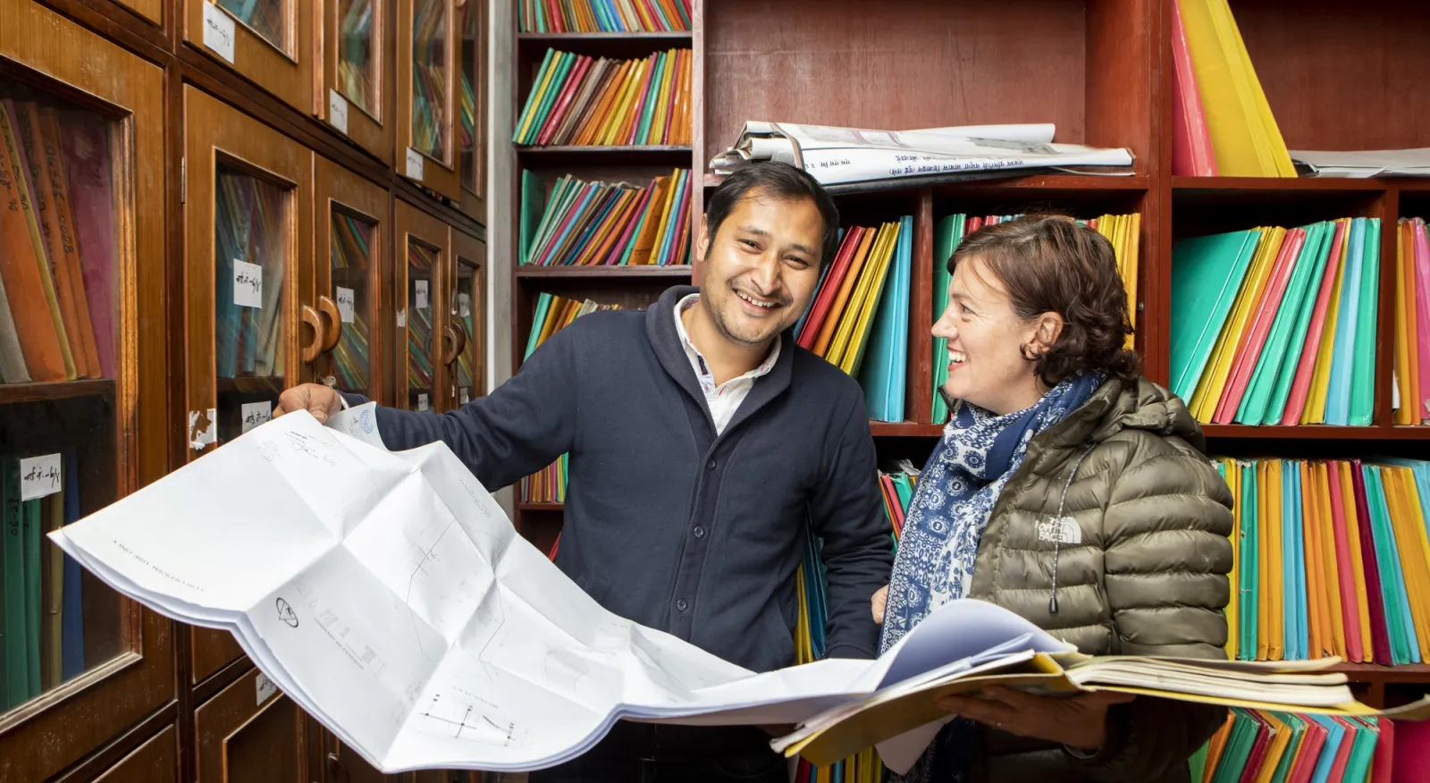 A man and a woman are dressed in jackets, standing in an office library filled with shelves of colorful folders. The man and woman have a large map open, and are smiling.