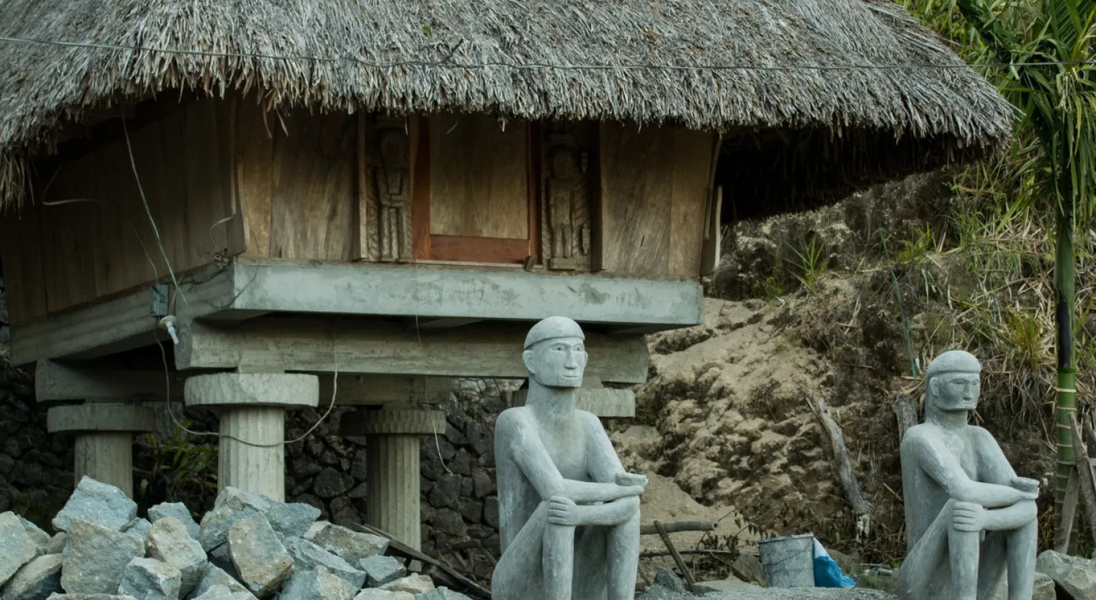 A scenic photo of a beachside building in the Philippines. There are statues of two men sitting outside the door, with rocks built up on either side.