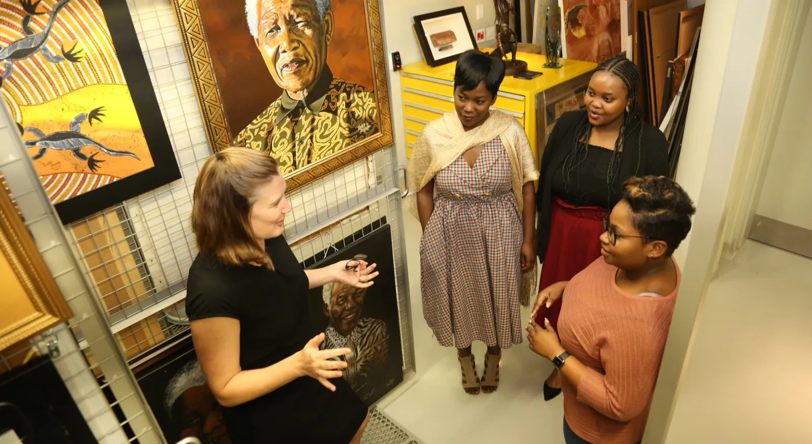 Four women stand in an art gallery talking, behind them is a large and prominent photo of Nelson Mandela.