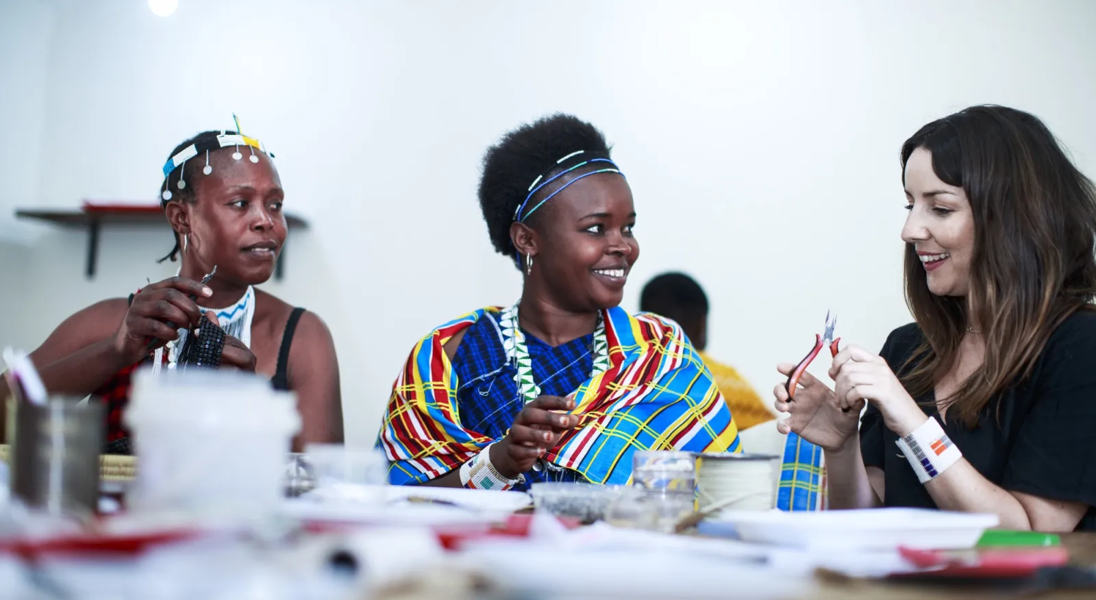 Three women sit at a table, beading jewlery. They are smiling and having a conversation.