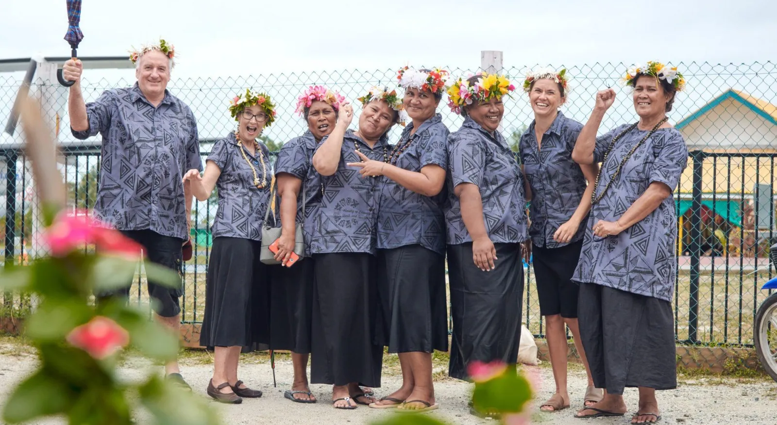 Seven women and one man all stand in a courtyard in matching printed shirts and flower crowns. They are grouped close together and smiling at the camera.