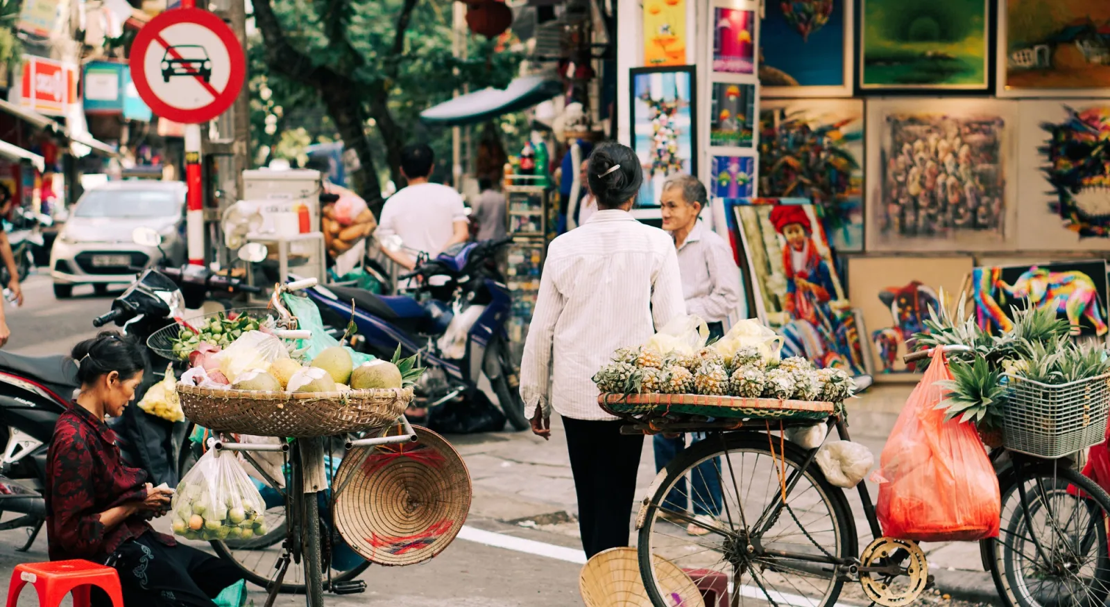 A woman walks across the street, away from the camera. She has a stack of fruit balanced on a wheeled cart. Another woman sits on the ground, selling food from her bicycle.
