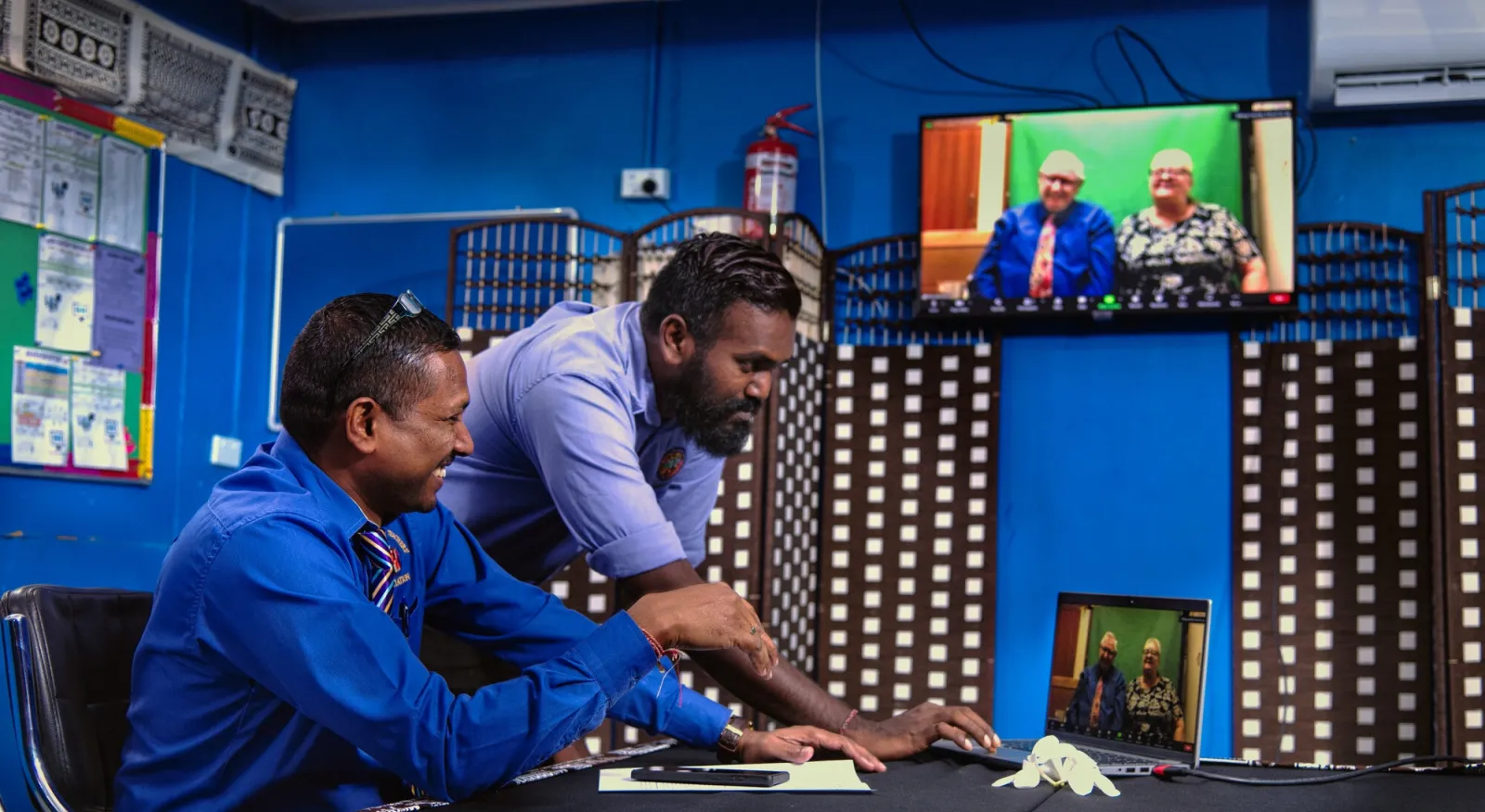 Two men sit at a desktop in a very blue room, working on a laptop. On the laptop a zoom screen is present with two people sitting and facing the camera.