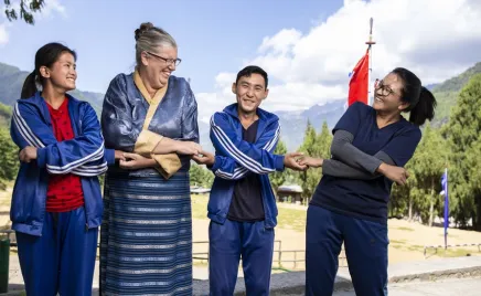 Four people in blue and red traditional Bhutanese clothing smiling and laughing while standing in front of a mountainous background.
