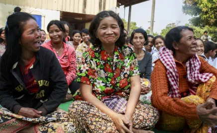 Three women sit on the ground in Cambodia, legs crossed and smiling in mid-conversation.
