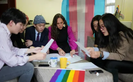 A group of young people are sitting around a small coffee table, with their heads down looking over papers. They are surrounded by a LGBTIQA+ flag and the Trans Right flag.
