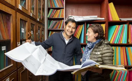 A man and a woman are dressed in jackets, standing in an office library filled with shelves of colorful folders. The man and woman have a large map open, and are smiling.