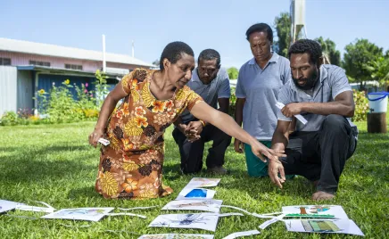 Four people are kneeling on grass, pointing at pieces of paper. They look to be mid-project.