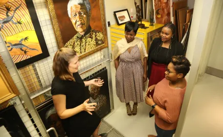 Four women stand in an art gallery talking, behind them is a large and prominent photo of Nelson Mandela.