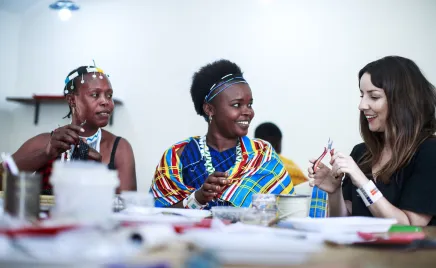 Three women sit at a table, beading jewlery. They are smiling and having a conversation.