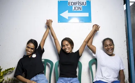 Three women sit in front of a directional sign. They clasp their hands together and lift their arms in the air while smiling and laughing.