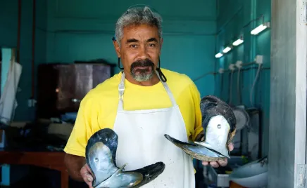A man in a yellow t-shirt and an apron hold two very large shells in his hands. He makes eye contact with the camera as he shows them off.