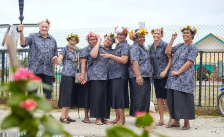 Seven women and one man all stand in a courtyard in matching printed shirts and flower crowns. They are grouped close together and smiling at the camera.