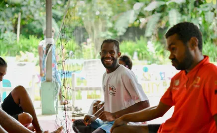 Two men sit on the ground with their legs crossed, in front of a volleyball net. One smiles at the camera, and the other is focused on the ongoing game.