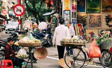 A woman walks across the street, away from the camera. She has a stack of fruit balanced on a wheeled cart. Another woman sits on the ground, selling food from her bicycle.