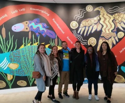 A group of six people stand in front of a large mural filled with Indigenous paintings of native animals. They are all smiling at the camera.