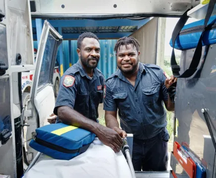 The camera is inside an ambulance, facing the back door where two men in Vanuatu paramedic uniforms standing outside smile at the camera.