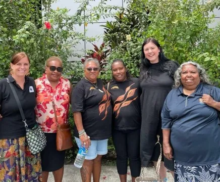 Six women stand outside a building and a lush garden in a tropical environment. They are all smiling, and some have arms wrapped around each other. Two of the women wear shirts with the Indigenous Pathways logo.