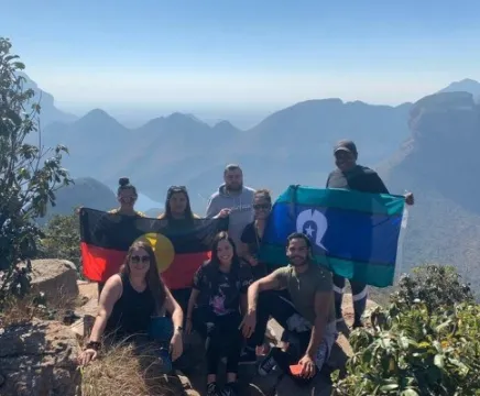 8 people stand on a mountain, with more peaks behind them. THey hold the Aboriginal and Torres Strait Islander flags in their arms.