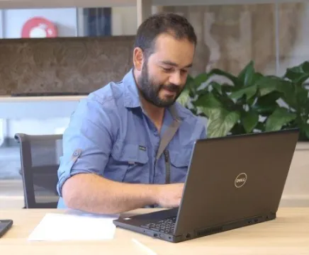 Clinton, a young man in a blue shirt, sits in front of a laptop in an office. He is typing and looking at the laptop screen.