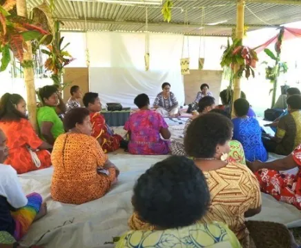 A group of women sit in a room together in Fiji, looking towards the presenters at the front of the room. All women are in colorful, floral dresses.