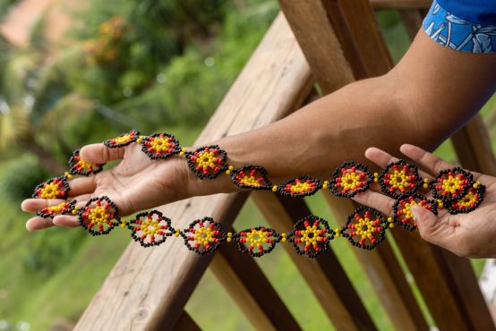 A beaded necklace done in black, red, and yellow. Hands display the necklace to the camera, with a lush garden in the background.