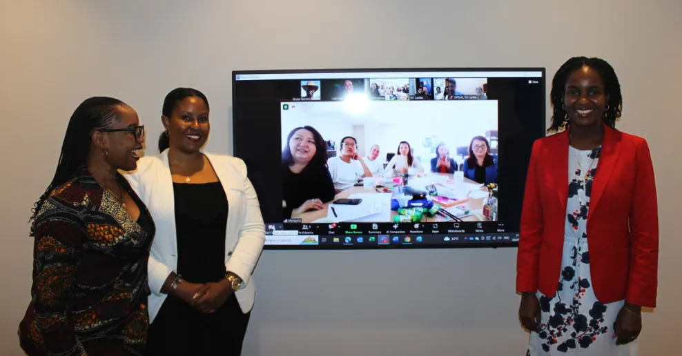 A screen with a team of people sitting around a table. Three people are standing on either side of the screen. Everyone in the image is smiling.