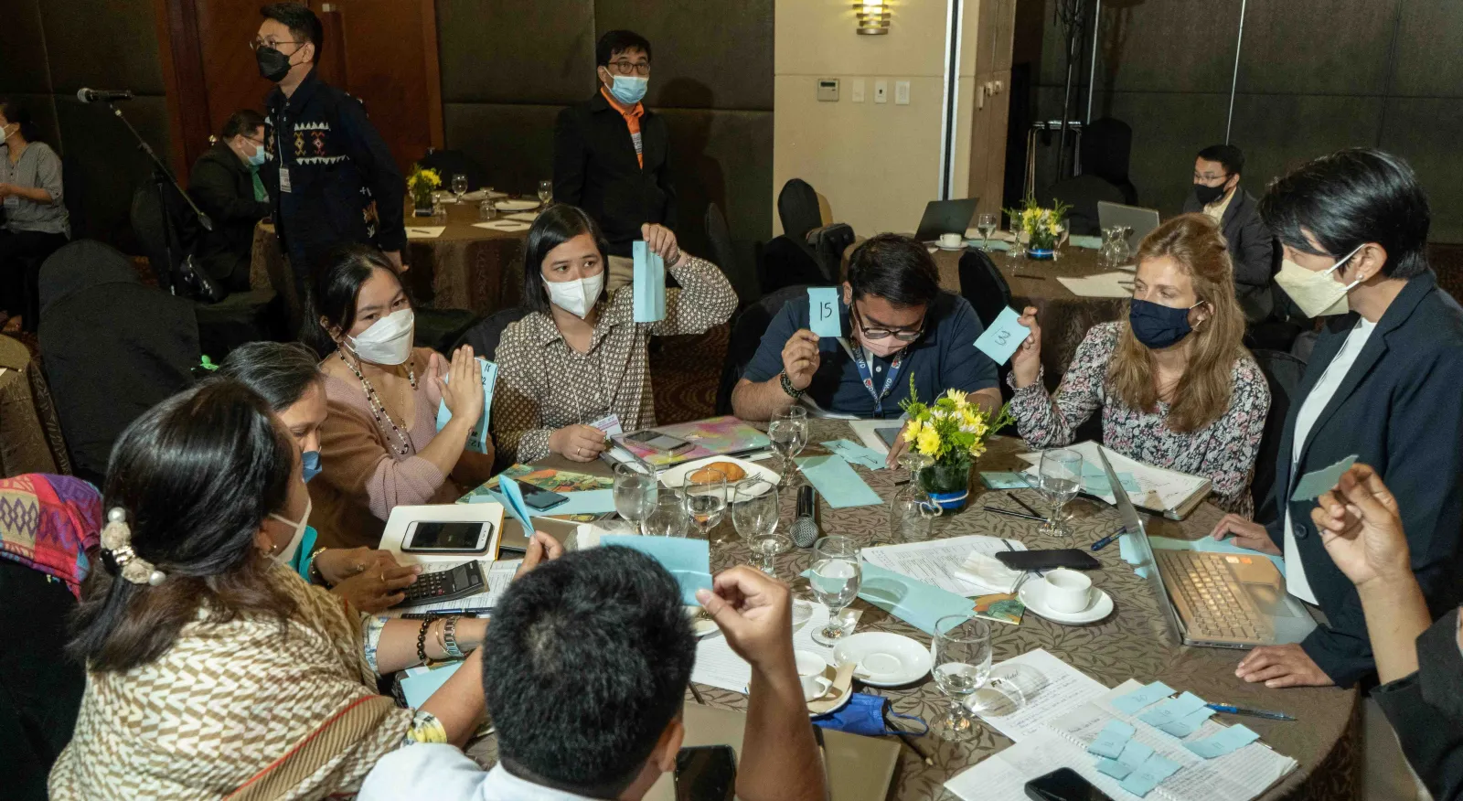 A group of people wearing masks are sitting around the table, actively working on a project together. The table is covered with paper and tea cups.