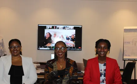 Three women in corporate attire sit in front of a Zoom screen showing people sitting around a desk, they all smile at the camera