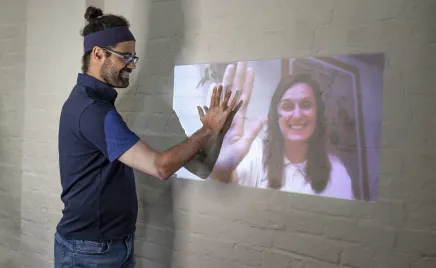 A man putting his hand on a screen display of a woman putting her hand in the same place.