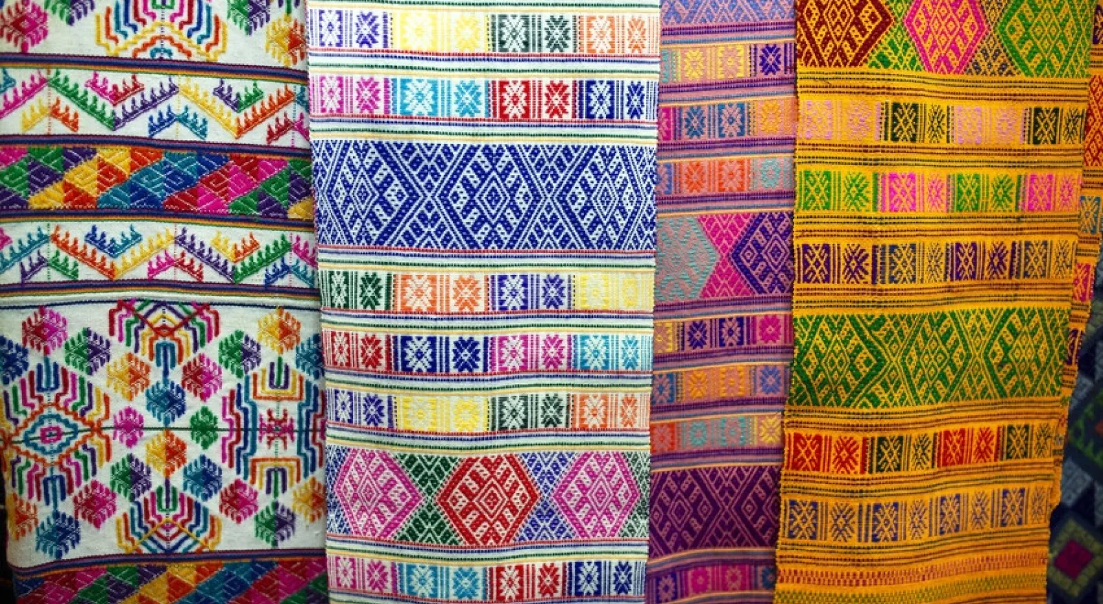 Four pieces of traditional woven fabric hang vertically, they are bright colours and very beautiful