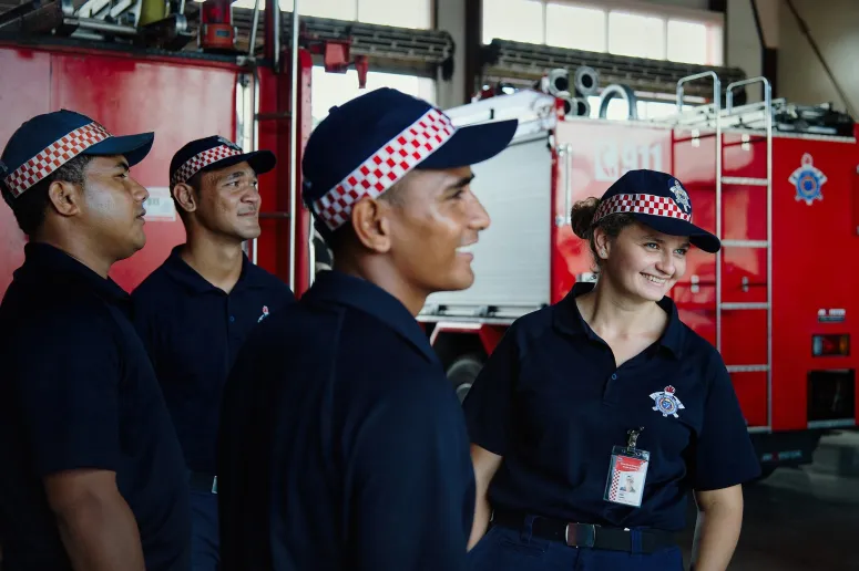 Three men and one woman wear firefighter uniforms and stand in front of a large fire truck