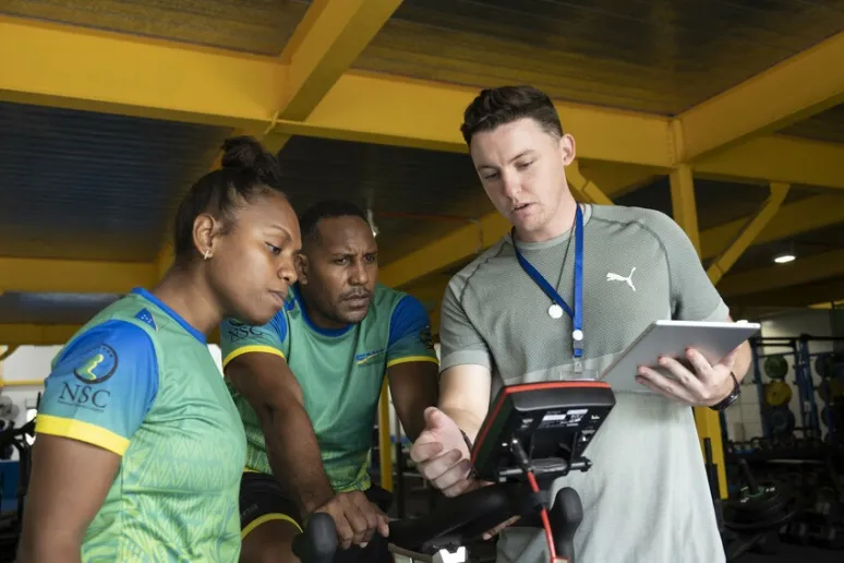 Three people in a gym. One is on an exercise bike following instructions from a volunteer. The third person is watching and listening over their shoulders.