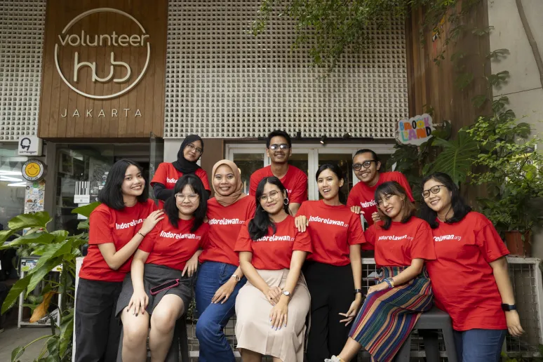 A group of 10 people stand outside a building that says Volunteer Hub Jakarta. They are all wearing matching red shirts that say Indorelawan, and are smiling at the camera.
