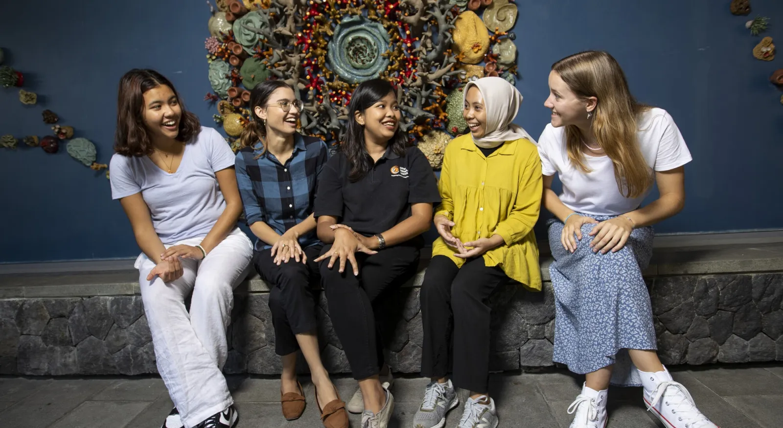 Four women sit on a bench chatting together, they sit in front of a sculpture