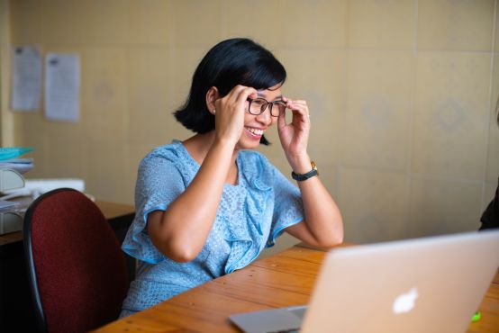 A young lady in a blue dress adjusts her glasses while sitting at a desk with a laptop on it