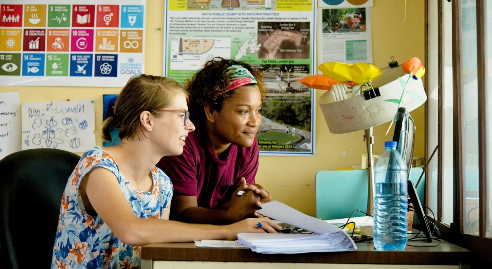 Two people sit at a desk, looking at a computer scream. The room is colorful, with plenty of posters and they look very engaged in their work.