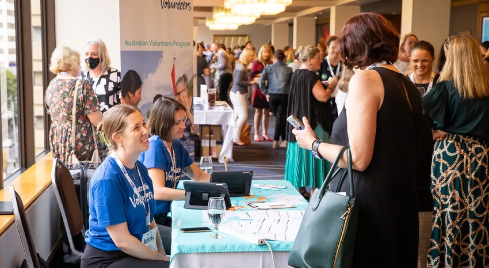 Two people sit at a conference table in a busy room. They wear matching Australian Volunteers t-shirts, and talk to another person who stands on the other side of the table.