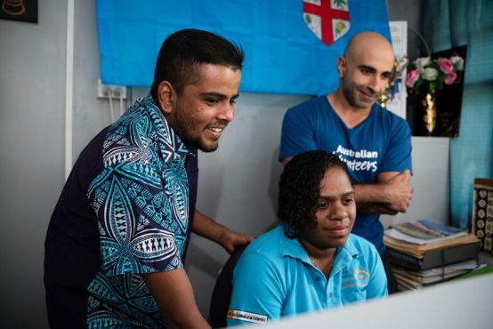 3 people working on one computer. One of the people is sitting at the desk with the other two supporting