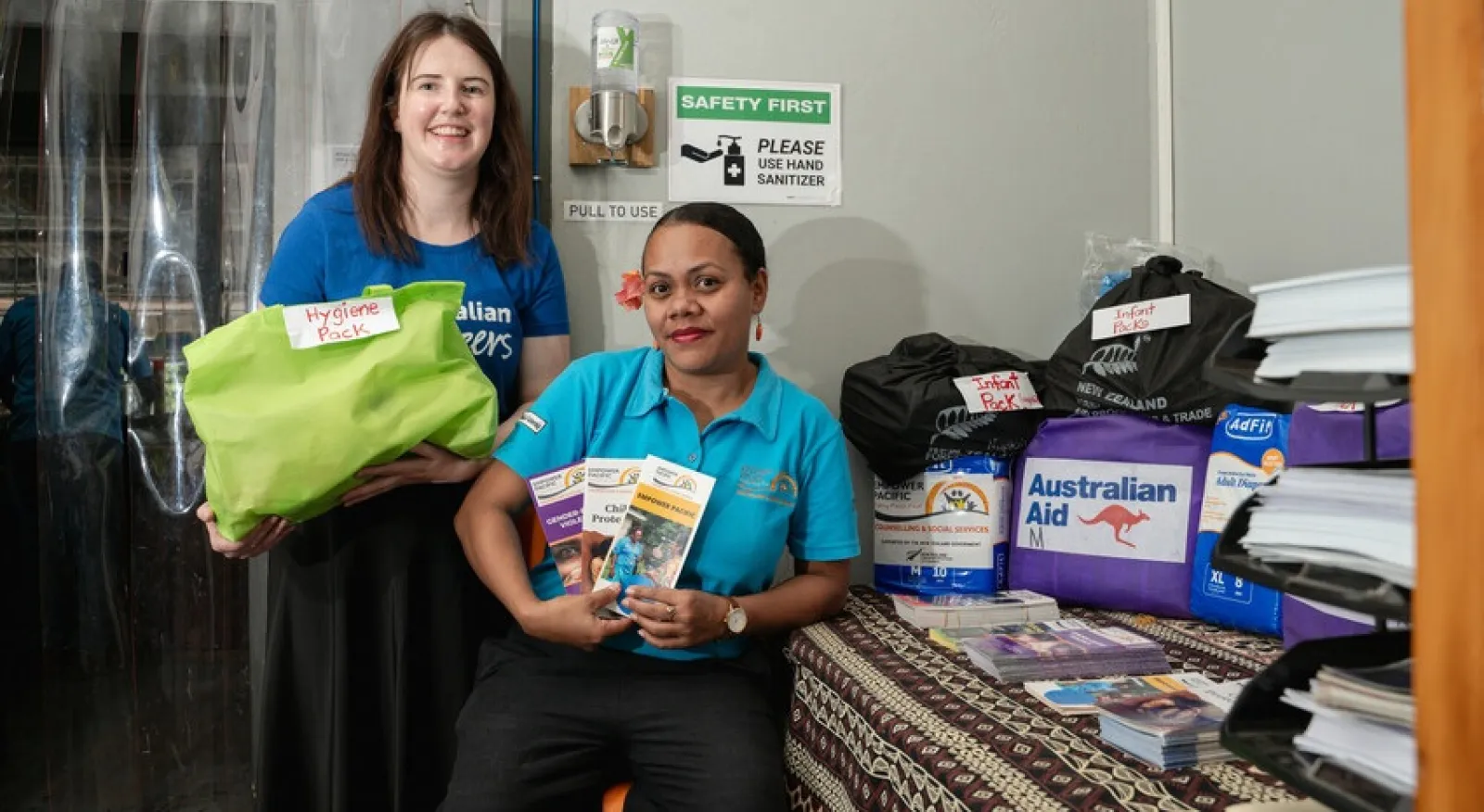 Two women smile directly into the camera. One stands and holds a package labelled 'hygiene pack' and the other sits on a stool while holding three brochures up. On the table beside them is a range of other collateral, including packages labelled 'Infant packs' and a range of brochures and leaflets.