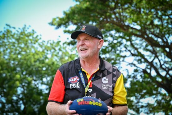 A man looks off into the distance with a candid expression on his face, while holding an AFL football in both his hands. He wears a cap, and t-shirt with the AFL PNG logos. There are trees and a blue sky in the background.