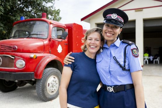 Two women stand together, smiling into the camera. They are standing outside a fire station, with a fire truck in the background, and the woman on the right is wearing a fire services uniform.
