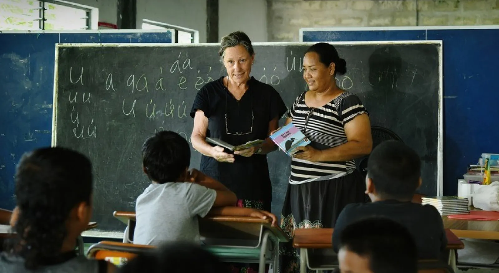 Two women stand at the front of a classroom. There is a blackboard behind them with chalk writing on it, and the women are captured candidly speaking to their students, who are seated at desks in front of them.